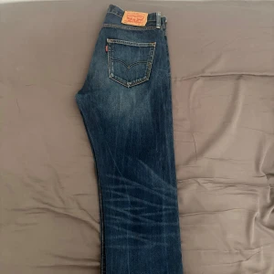 Levi's 501 blå jeans straight fit - Klassiska Levi's 501 jeans i blå denim med snyggt slitna detaljer och raka ben. Jeansen har fem fickor, knäppning med knappar och den ikoniska läderpatchen bak i midjan. Perfekta för dig som gillar tidlös stil och vill ha ett par jeans som alltid funkar. Storlek 32/34