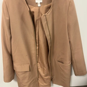 Beige kappa från H&M med dragkedja - Stilren beige kappa från H&M med rak passform och diskret dragkedja framtill. Jackan har två fickor med lock och är helfodrad. Perfekt för dig som gillar en clean och klassisk look.