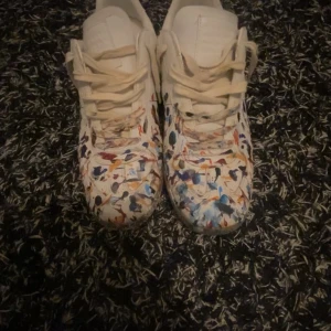 Maison Margiela sneakers med färgstänk - Unika vita sneakers från Maison Margiela med coolt färgstänkt mönster i blått, rött, gult och svart. Klassisk rund tå, snörning och platt sula. Skorna är i skinn och har en riktigt konstnärlig vibe som sticker ut.