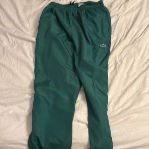 Gröna byxor från Lacoste med resår - Snygga gröna byxor från Lacoste med klassisk krokodillogga på benet. Byxorna har resår i midjan och är gjorda i ett glansigt syntetmaterial. Perfekta för en chill och sportig look.