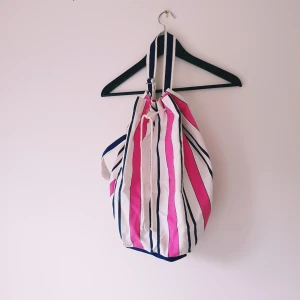 Randig ryggsäck - Säljer en stor och mjuk ryggsäck i bomull med breda rosa, vita och mörkblå ränder. Väskan har dragsko upptill och en bred axelrem i samma mönster. Perfekt för att slänga ner allt du behöver till stranden eller stan.  Helt skick dock obehandlade fläckar