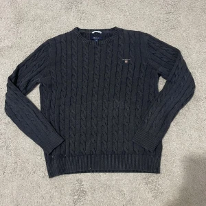 Mörkgrå kabelstickad tröja från GANT - Kabelstickad mörkgrå tröja från GANT med rund hals och broderad logga på bröstet. Tröjan har ribbade muddar vid ärmslut och nederkant, samt långärmad! Är i storlek M men sitter som S.