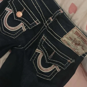True Religion Becca Bootcut Jeans - Mörkblå bootcut jeans från True Religion, modell Becca. Snygga kontrastsömmar i vitt och rosa, klassiska fickor med broderad logga och kopparfärgade knappar.  De har heelbites men annars i bra kondition, tryck på köp nu💗