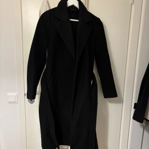 Svart lång kappa med bälte - Svart kappa med bälte i midjan. Från VERO MODA , köptes för 699kr och knappt använd. Har inga skador !🤍är i storlek XS men passar en med S också💕