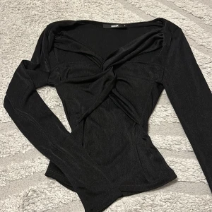 Svart omlottopp från BikBok - Säljer en svart långärmad omlottopp från BikBok. Toppen har en snygg twistad detalj framtill och v-ringning som ger en cool och trendig look. Materialet är mjukt och stretchigt, perfekt att styla med jeans eller kjol.