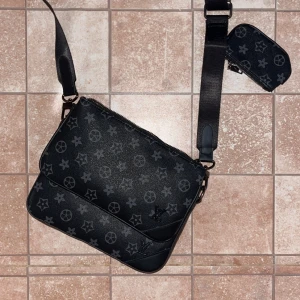 Helt ny svart axel väska - Helt ny. Snygg svart axelväska med klassiskt monogrammönster i grått. Väskan har justerbar axelrem, flera fack med dragkedja och en liten avtagbar plånbok. Tillverkad i skinn med detaljer i svart och grått. Perfekt för dig som vill ha en stilren och lyxig look.