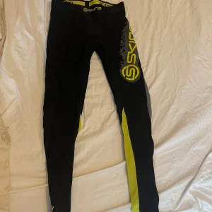 Svarta Skins träningstights S - Svarta träningstights från Skins med gula och grå detaljer samt stor logga på benet. Tightsen har elastisk midja med Skins-tryck och är tillverkade i ett stretchigt syntetmaterial som sitter tight mot kroppen. Perfekta för löpning eller gym.