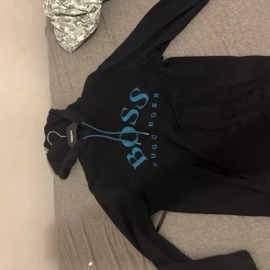 Svart hoodie från Hugo Boss med blå logga - Snygg svart hoodie från Hugo Boss med stor blå BOSS-logga framtill och matchande blå snören i huvan. Klassisk passform med långa ärmar och mjukt material, perfekt för en chill och stilren look.