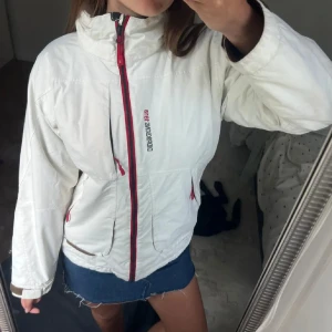 Vit vindjacka från Didriksons 1913 - Snygg vit vindjacka från Didriksons 1913 med röda detaljer och dragkedjor. Jackan har hög krage, justerbara ärmslut och flera fickor framtill. Perfekt för blåsiga dagar och enkel att matcha med olika outfits.