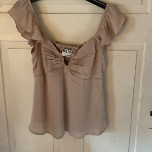 Beige blus med volangärm från Aware - Supersöt beige blus från Aware med volangärmar och smockad rygg. Toppen har snygg rynkdetalj framtill med silvriga knappar. Sitter tajtare vid bröstet och luftigare nedtill. Använts en gång. 