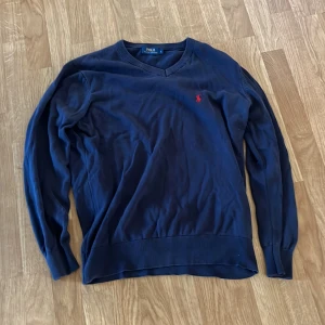 Mörkblå v-ringad tröja Polo Ralph Lauren - Mörkblå v-ringad tröja från Polo Ralph Lauren i storlek S. Tröjan har lång ärm, ribbade muddar och en röd broderad logga på bröstet. Perfekt för dig som gillar klassisk och stilren design.
