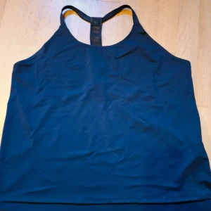 Svart träningstopp Fabletics Demi Lovato - Snygg svart linnetopp från Fabletics Demi Lovato-kollektionen. Modellen har smala axelband och meshdetalj i ryggen för extra ventilation. Perfekt för träning och tillverkad i stretchig polyesterblandning som sitter skönt på kroppen.