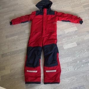Röd overall från Didriksons - Säljer en röd och svart overall från Didriksons med huva, dragkedja och tryckknappar. Overallen har flera fickor, förstärkta knän och reflexdetaljer vid benen. Perfekt för kalla dagar och vinteraktiviteter. Materialet är slitstark syntet. Använd 2 gånger