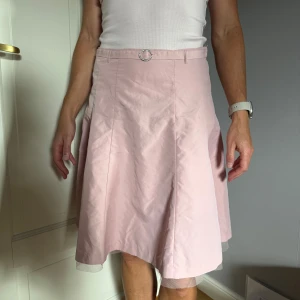 Ljusrosa A-linjeformad kjol - Supersöt ljusrosa kjol med A-linjeform och diskret glans. Kjolen har en liten dekorativ strassprytt skärp och ett tunt lager tyll längst ner som sticker ut. Perfekt för dig som gillar en feminin och stilren look.