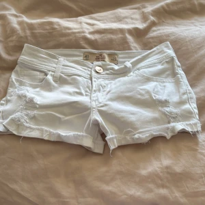 Vita low rise jeansshorts Hollister - Snygga vita jeansshorts från Hollister med låg midja och råa, fransiga kanter. Shortsen har slitna detaljer framtill, klassiska fickor och knapp med Hollister-logga. Perfekta för varma dagar när du vill ha en chill och trendig look.
