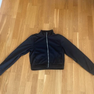 Svart träningsjacka från SOC, 158/164 - Svart träningsjacka från SOC i sportig modell med hög krage och hel dragkedja framtill. Jackan har långa ärmar samt ribbad nedkanter. Perfekt för dig som gillar en clean och enkel stil.