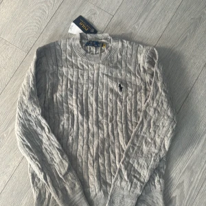 Grå kabelstickad tröja Polo Ralph Lauren - Kabelstickad grå tröja från Polo Ralph Lauren med klassisk rund halsringning och broderad logga på bröstet. Mjuk och skön, perfekt för lager-på-lager. Långärmad och ribbade muddar för en clean look.