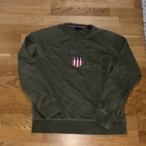 Olivgrön sweatshirt från GANT - Olivgrön sweatshirt från GANT med broderad logga och sköld på bröstet. Tröjan har rund halsringning, långa ärmar och ribbade muddar. Klassisk och stilren design med inslag av rött, vitt och blått i detaljen på insidan av kragen.