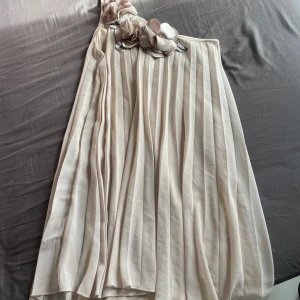 Kort gullig klänning - Elegant champagnefärgad klänning från Elise Ryan med vackra blommiga detaljer vid axeln. Klänningen har en oneshoulder-design och ett plisserat tyg som ger en fin struktur. Perfekt för sommartillfällen eller skolavslutning!