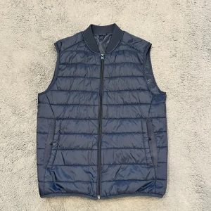 Mörkblå dunväst från Zara - Snygg mörkblå dunväst från Zara med dragkedja och två fickor framtill. Perfekt för lager-på-lager under kyliga dagar. Västen har en stilren design med ribbad krage. Storlek s