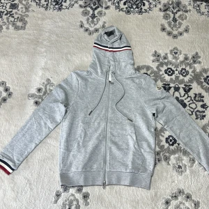 Grå huvtröja från Moncler - Säljer en stilren grå huvtröja från Moncler med dragkedja och ribbade muddar. Tröjan har en justerbar huva med röd och vit randdetalj samt Moncler-logga på ärmen. Perfekt för en avslappnad look.