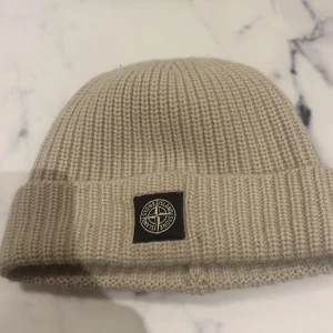 Beige stickad mössa från Stone Island - Snygg beige stickad mössa från Stone Island med uppvikt kant och logotyp framtill. Perfekt för att hålla värmen under kyliga dagar. En stilren accessoar som passar till det mesta.