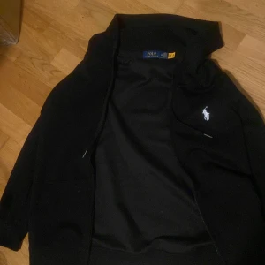 Polo Ralph Lauren zip hoodie - Säljer en stilren svart zip hoodie från Polo Ralph Lauren med dragkedja och broderad logga på bröstet. Den har en klassisk design som passar perfekt för både höst och vår. Det är en storlek M/Medium och den är helt ny/Aldrig använd. Pris kan diskuteras✅