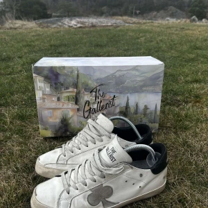 Vita sneakers från The Gallerist - Snygga vita sneakers från The Gallerist med svart häl och ett stort klövermotiv på sidan. Skorna har snörning och en rund tå, perfekt för en stilren look.