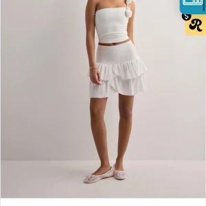 Vit volangkjol neo noir - Söt vit kortkjol med volanger och smockdetaljer i midjan. Perfekt för sommarens varma dagar. Kjolen har en luftig och lekfull design som ger en romantisk touch.