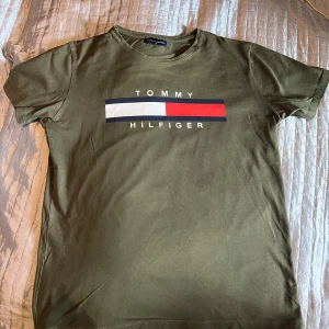 Olivgrön t-shirt från Tommy Hilfiger - Säljer en olivgrön t-shirt från Tommy Hilfiger med deras ikoniska logga i vitt, rött och blått på bröstet. T-shirten är kortärmad och gjord i mjuk bomull. Perfekt för en avslappnad stil. Den är dock mindre i storleken och passar som M