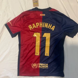 Blå och röd fotbollströja från Nike Barcelona  - Säljer en snygg fotbollströja från Nike i blått och rött med korta ärmar. Tröjan har ett tryck med 'Raphinha 11' på baksidan och FC Barcelona-loggan på framsidan. Perfekt för fotbollsfans!