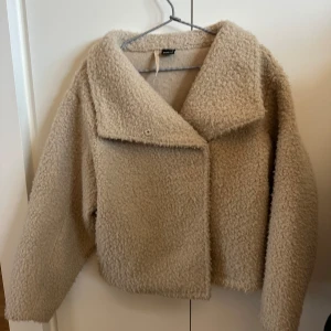 Beige teddyjacka från Ginatricot - Mysig beige teddyjacka från Ginatricot med en oversized passform. Jackan har långa ärmar och en stor krage. Den stängs med knappar framtill och har en mjuk och fluffig textur.