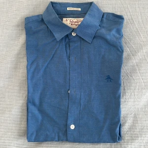Blå skjorta från Original Penguin - Snygg blå skjorta från Original Penguin med klassisk krage och knappar framtill. Skjortan har en diskret broderad logga på bröstet och är tillverkad i ett lätt och bekvämt material.