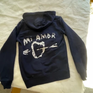 Mörkblå hoodie med tryck - Säljer en mörkblå hoodie med ett coolt tryck på från mira Paris ,på baksidan står de  'Mi Amor' med en pil genom ett hjärta. Den har en klassisk känguruficka och en bekväm huva. Perfekt för en avslappnad stil. OBS : den har ett litet hål , se sista bilden . Ny pris 1500kr mitt pris 650kr, upplagd på flera sidor så va snabb om du vill köpa!💕