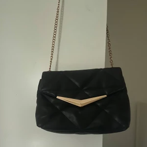 Svart quiltad axelväska med kedja - Snygg svart axelväska med quiltat mönster och guldfärgad kedja. Väskan har en elegant design med en dekorativ guldfärgad detalj på framsidan. Perfekt för att ge din outfit en stilfull touch.