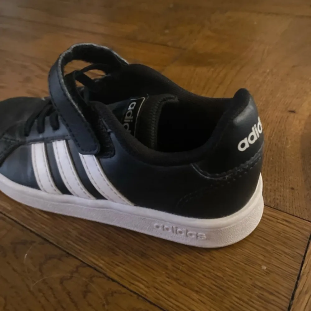 Snygga svarta sneakers från Adidas med klassiska vita ränder och vit sula. Skorna har snörning och kardborreband för extra säker passform. Perfekta för vardagsbruk och sportiga aktiviteter.. Kengät.