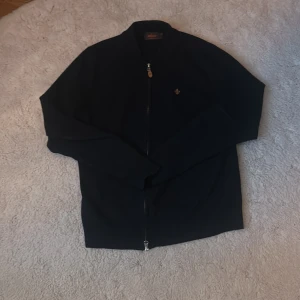 Marinblå fullzip tröja från Morris - Snygg marinblå fullzip tröja från Morris i 100% merinoull. Den har en dragkedja framtill och en diskret broderad logotyp på bröstet. 