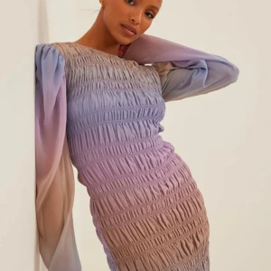 Lila klänning från NA-KD - Säljer en vacker lila klänning från NA-KD med långa ärmar och ett smockat mönster. Klänningen har en mjuk övergång mellan olika lila nyanser och är perfekt för en stilfull look. Den är tillverkad av ett lätt och luftigt material. Helt ny med prislapp. Se sista bilden för en väldigt smuts fläck. 