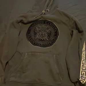 Svart hoodie från Glorious Gangsta - Säljer en svart hoodie från Glorious Gangsta med ett stort emblem på bröstet och dekorativa mönster längs ärmarna. Hoodien har en klassisk känguruficka och justerbar huva med snören. Perfekt för en stilren och avslappnad look.