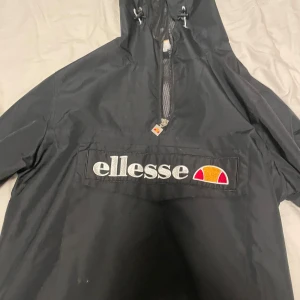 Svart vindjacka från Ellesse - Säljer en svart vindjacka från Ellesse med dragkedja och huva. Jackan har ett stort Ellesse-märke på framsidan och en broderad patch med texten 'Cortina D'Ampezzo' på ärmen. Perfekt för kyliga dagar och sportiga aktiviteter.