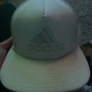 Vit keps från Adidas - Snygg vit keps från Adidas med broderad logotyp framtill. Perfekt för en sportig look eller sommarens äventyr. Kepsen har en klassisk design med böjd skärm.