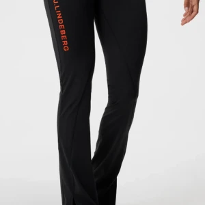 Svarta byxor från J.Lindeberg - Snygga svarta leggings byxor från J.Lindeberg med orange logotyp längs benet. Byxorna har en dragkedja framtill och är tillverkade i ett syntetmaterial som ger en bekväm passform. Perfekta för en stilren look, slit längst nere