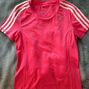 Rosa träningstopp från Adidas - Säljer en snygg rosa träningstopp från Adidas med de klassiska tre ränderna i vitt på ärmarna. Toppen är kortärmad och tillverkad i ett lätt och andningsbart material, perfekt för träning. Adidas-loggan är tryckt på bröstet. Passar perfekt för dig som vill träna med stil! 💪