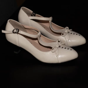  pumps med rosett från Ecosusi - Vintage Pumps från Ecosusi Vintage i modern vintage design. De förtrollande skorna med liten klack och spetsig form är dekorerade med en dekorativ rosett på framsidan, som understryker vintagekaraktären. De justerbara remmarna hakar fast i sidan, vilket gör den lätt att ta på.  Gjorda av vegansk konstläder. Skorna är använda en gång på en väns bröllop. Orginalpris är ca 800 kr.  Klackhöjd: 5 cm 
