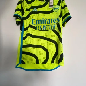 Gul och svart Arsenal fotbollströja - Säljer en cool gul och svart Arsenal fotbollströja från Adidas. Tröjan har korta ärmar och ett unikt mönster med svarta linjer och prickar. Den har klubbens logga och sponsortryck på framsidan. Perfekt för alla Arsenal-fans! Storlek M passar L