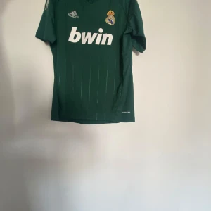 Grön Real Madrid fotbollströja - Säljer en grön Real Madrid fotbollströja från Adidas med korta ärmar. Tröjan har klubbens emblem och sponsorlogga på framsidan samt spelarnamn och nummer på baksidan. Perfekt för fans som vill visa sitt stöd! Stl M kan passa L