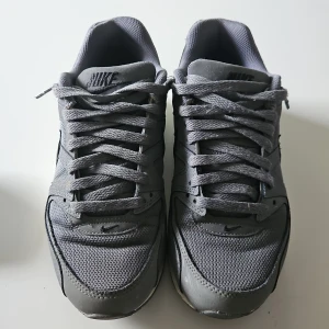 Grå Nike Air Max sneakers - Snygga grå Nike Air Max sneakers med klassisk design och synlig luftdämpning i sulan. Skorna har snörning och är tillverkade i syntetmaterial med meshdetaljer för extra andningsförmåga. Perfekta för både vardag och träning.