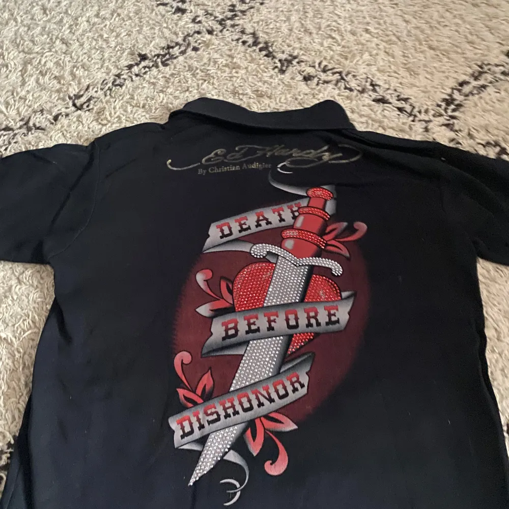 Säljer en svart kortärmad t-shirt från Ed Hardy med ett stort tryck på ryggen som säger 'Death Before Dishonor'. T-shirten har en klassisk krage och är gjord i bomull. Perfekt för dig som gillar en edgy stil!. T-paidat.