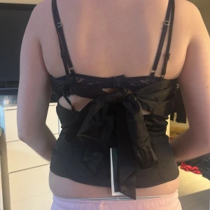Svart korsettaktig topp med rosett - Snygg svart korsett med justerbara axelband och en stor rosett i ryggen. Toppen har en elegant v-ringning och en dragkedja på sidan för enkel påklädning. Perfekt för en stilfull look.