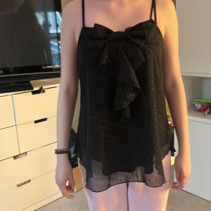 Svart ärmlös topp från Vila - Säljer en elegant svart ärmlös topp från Vila med tunna axelband och en stor rosett framtill. Toppen har ett subtilt prickigt mönster och en volangdetalj som ger en extra touch. Perfekt för en stilren look. Det prickiga mönstret är lite glittrigt.